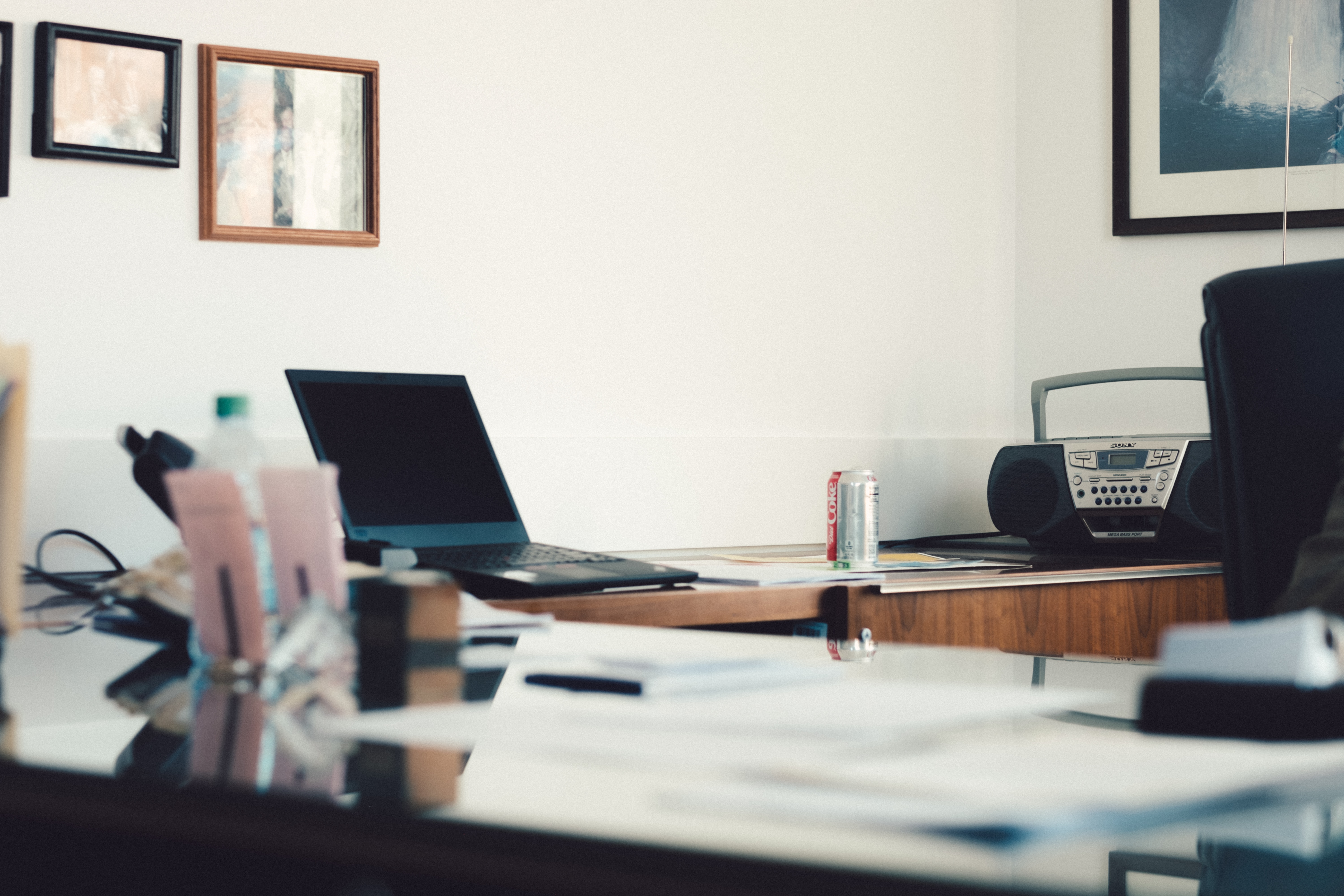 The image depicts an office desk in a workspace. The desk is cluttered with various items, including a closed laptop, office supplies, papers, and a can of Diet Coke. A stereo system is positioned on the wooden desk surface against the wall. The desk is shiny, partially reflecting some objects. The wall behind the desk is adorned with framed pictures; one has a wooden frame, and two others have black frames. The office chair is partially visible on the right side of the image. 