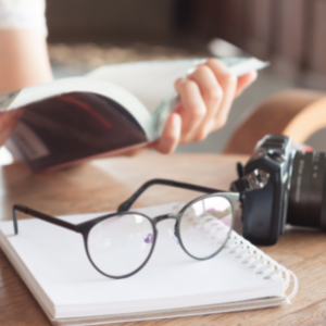 Pair glasses sit on a notebook on a desk. In background is a persons hands reading a book.