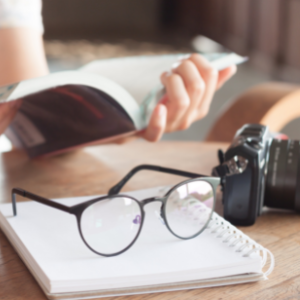 Une paire de lunettes est posée sur un cahier sur un bureau. En arrière-plan, on voit les mains d'une personne qui lit un livre.