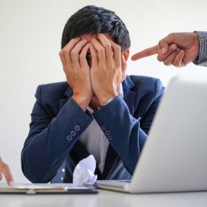 Homme assis à un bureau d'ordinateur, les mains couvrant son visage. Une autre personne pointe son doigt vers sa tête en signe de désapprobation.