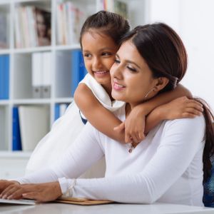 Mother working at computer with her young daughter hugging her neck from behind. 