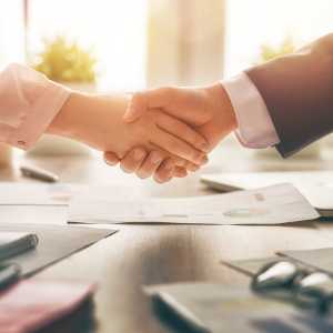 Two people shaking hands over a table with documents in an office setting.