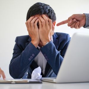 Male sitting at computer desk with hands covering his face. Another persons finger is pointed at his head as in disaproval.